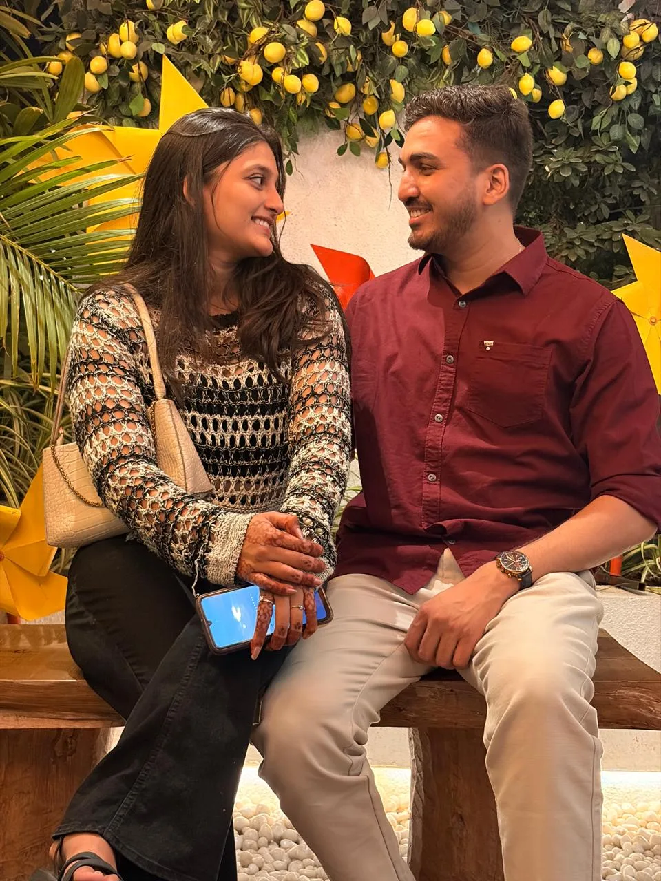 Neha and Sahil on a bench, smiling at each other beneath a wall of lemons